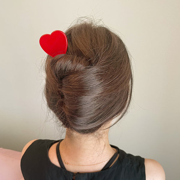 Acetate hair stick with a heart shaped topper in a shade of bright red. Shown in a models hair from the back 