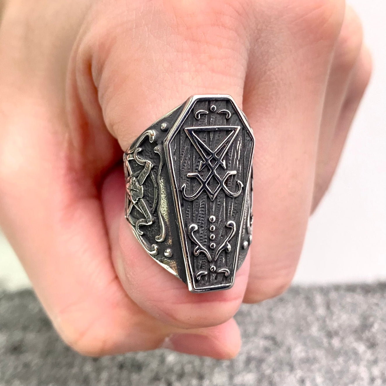 A stainless steel ring with dark antiqued detailing in the shape of a coffin decorated with various occult symbols. Shown worn on a pointer finger in a fist