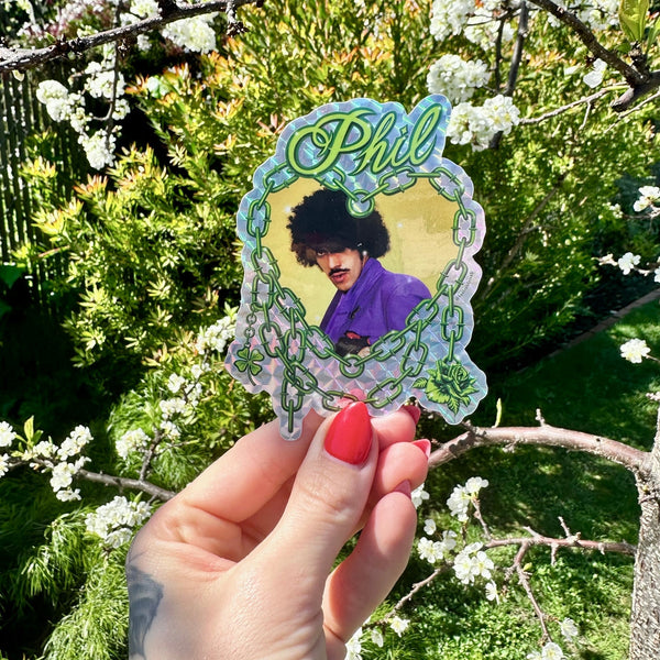 Holographic die-cut vinyl sticker featuring a portrait of Phil Lynott from Thin Lizzy wearing a purple shirt on a yellow background inside of an illustration of a neon green chain link heart with “Phil” in green script. Shown held in front of a floral background