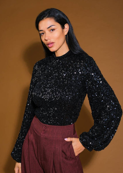 model wearing a black top with high neckline and long balloon-style sleeves in an allover black sequin fabric. Shown from front