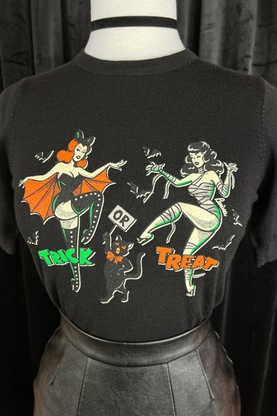 black crew neck short sleeved sweater with retro women wearing bat costume and mummy costume alongside a black cat with message "TRICK OR TREAT". Shown on dress form in close up tucked into leather skirt