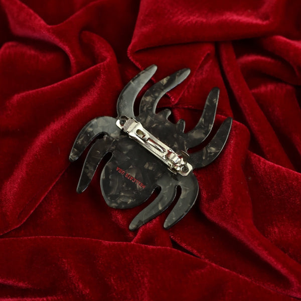 black marbled acetate hair clip in the shape of a spider with red and pink line detail and pink eyes. Shown from back on red velvet background