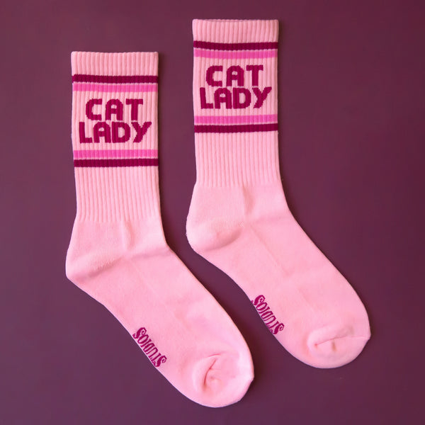light pink cotton crew socks with ribbed cuffs and 'CAT LADY' text in magenta between pink stripes. Shown flat on a purple background