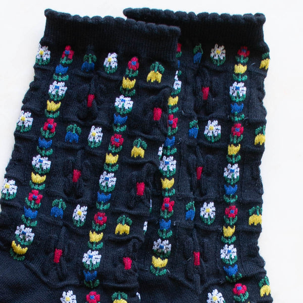 cotton knit socks in black with an allover red, yellow, green, blue, and white daisy and tulip knit-in pattern. Shown in close up 