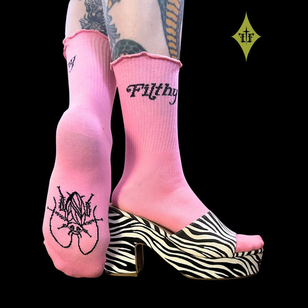 Bright pink cotton-poly blend crew socks with lettuce hem-style ruffled ribbed cuffs and “Filthy” written in black script running across each outer cuff and black outline cockroach on bottom of sock. Shown worn by model wearing platform zebra print mule sandals with one foot raised to show bottom of sock and side of sock