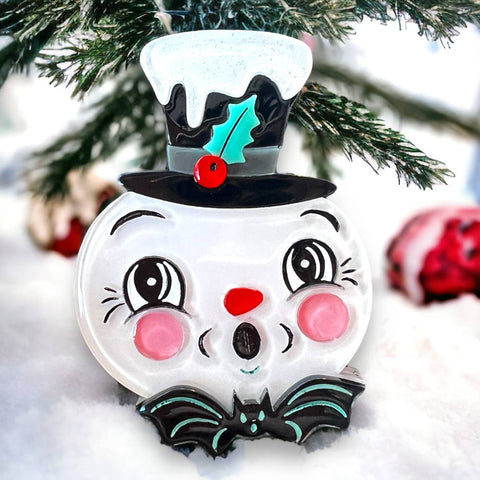 Spooky Frosty Brooch by Johanna Parker x Lipstick & Chrome, surprised face snowman wearing top hat covered with white glittery snow and holly detail with small bat at his collar. Shown on a snowy tree background 