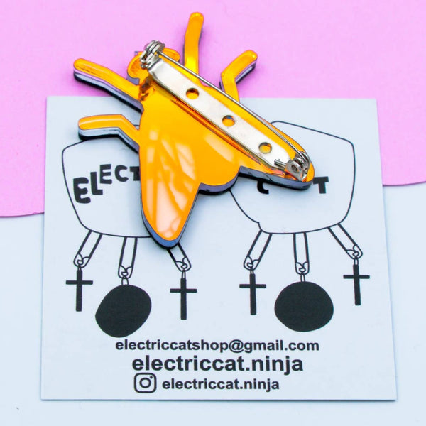 layered acrylic brooch of a black fly with iridescent translucent blue wings. Shown on backing card displaying brooch pin hardware