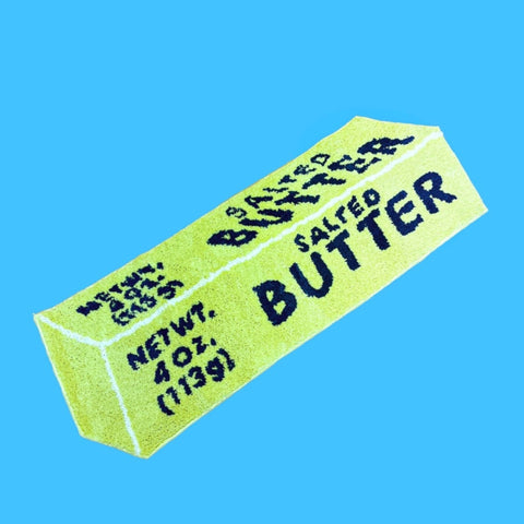 Rug shaped like a yellow stick of butter with blue lettering and a white outline. Shown flat on a blue background 