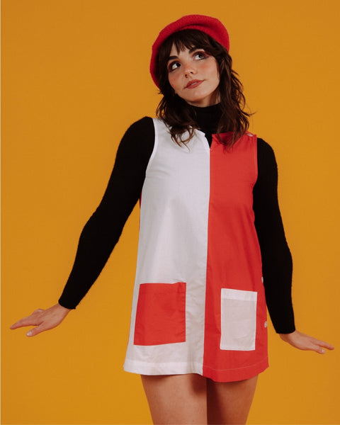 a model wearing a cotton a-line smock style mini dress in half red, half white design divided vertically down the middle of the garment. it has a round neckline with slash detail, button closure running down the left shoulder and down the side of the dress, and patch pockets in alternate colors at each hip. Shown from front styled with black turtleneck and red beret