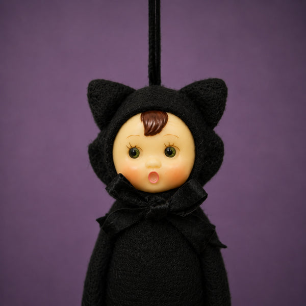 Poseable ornament with wire limbs and Velcro hands in the shape of a pixie with rosy cheeked, green eyed resin face with molded tuft of brown hair. She is wearing a black faux fur black cat costume with ears and tail. Black satin ribbon for hanging is at top of head. Shown in close up of face on purple background