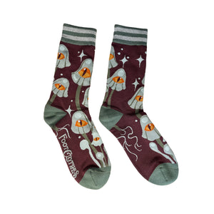 Unisex crew socks with an all-over pattern of green-grey mushrooms each with a large yellow and orange eye on their caps. On a purple-ish brown background surrounded by matching grey starbursts. Striped green and grey cuffs and solid green toes and heels. Seen flat