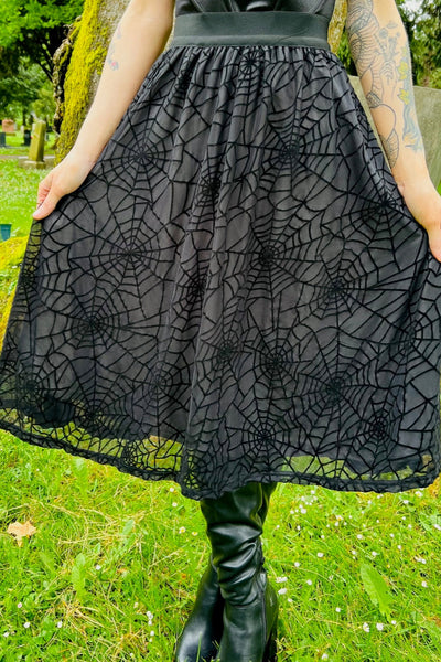 Model wearing a high waisted black below the knee length full skirt with a 2” wide elastic waistband, outer layer of sheer black mesh with a black flocked spiderweb design, and inner lining. Shown from the front with skirt held taut