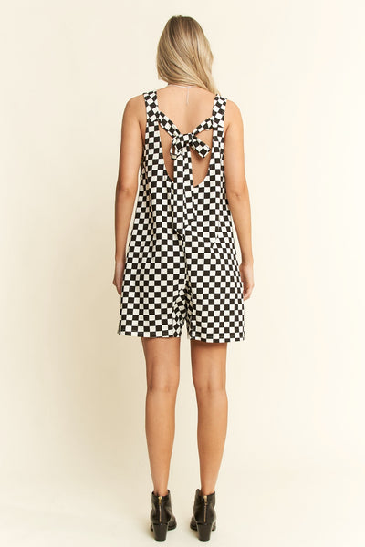 model wearing a black and white checker print cotton blend relaxed fit romper with wide straps, slightly rounded v-neckline, self-tie detail at back, relaxed fit shorts with patch pockets. Shown from back