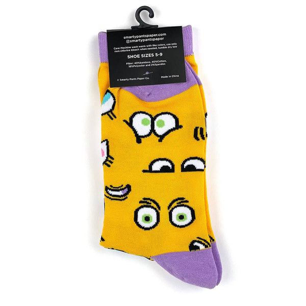 Crew socks with sets of cartoon eyes on a golden yellow background with lavender cuffs, heels, and toes. Shown flat in packaging from back