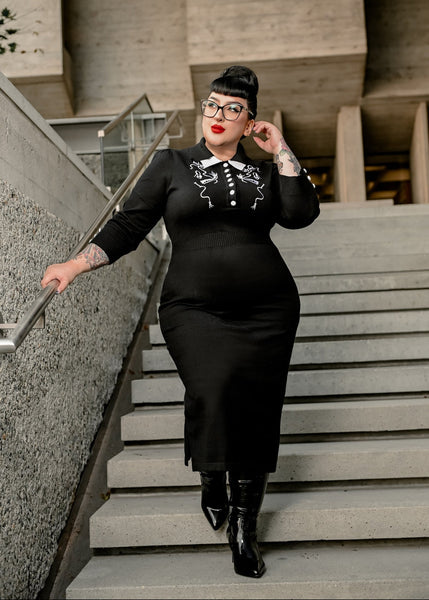 Plus size model wearing a black medium-weight knit sweater dress with double layered collar in black and white, bodice with white button-down front and cuffs, ribbed waistband and elbow-length sleeves, and mirrored embroidered detail of an opera-length glove, kitten heel, and coiled whip. Skirt is pencil length with double side slits. Shown from the front