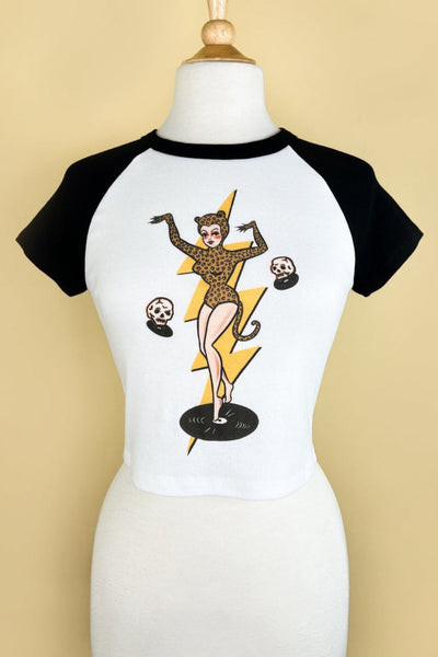 white crop t-shirt with black raglan sleeves & collar and printed illustration of woman wearing leopard print catsuit with ears and tail posing on a vinyl record in front of a yellow lightning bolt and surrounded by skulls. Shown on dress form