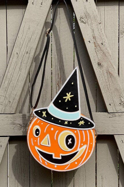 Faux leather shoulder bag in the shape of an orange jack-o’-lantern pumpkin with smiling face and black witch hat with yellow stars. Bag has adjustable black faux leather strap. Shown from front hanging on fence