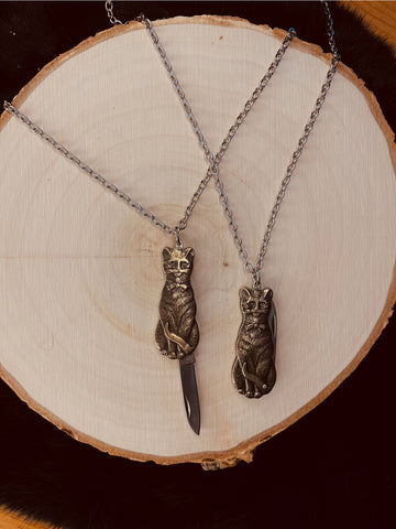 Mini pocket knife charm in the shape of a cat with antiqued brass finish. On a 20” silver metal link chain. Shown flat and open next to a second necklace with knife closed on a tree stump.