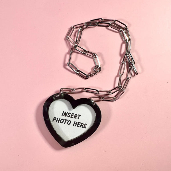 A heart-shaped marbled black pendant necklace with a white center that reads 'INSERT PHOTO HERE', connected to a silver paperclip chain on a solid pink background.