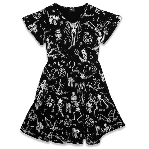 black cotton jersey knit dress with white skeleton, owl, pumpkin, witch and bat print. Dress has a shallow v-neckline, short flutter sleeves, a knee length double tiered skirt with ruffle hem and pockets. Shown from front on white background 