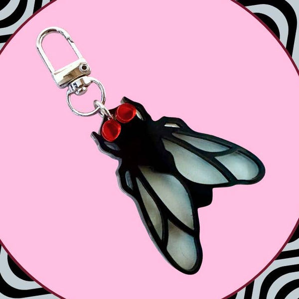 Laser-cut acrylic in the shape of a house fly with black body and transparent matte finish blue wings and red mirrored eyes. Attached to silver metal keychain via silver metal O-ring. Shown on pink background