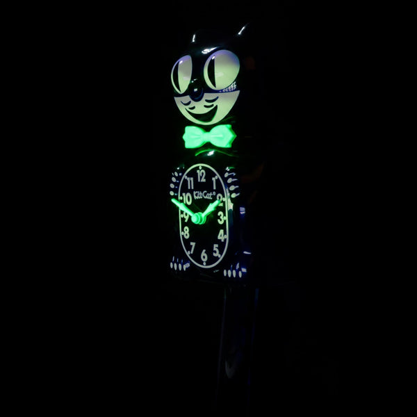 black & white glow in the dark Kit-Cat wall mount clock features a mischievous grin, and big round eyes that swivel side-to-side in time with its pendulum tail. Shown in dark with glowing bowtie and clock hands from three quarter angle