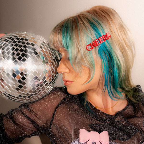 Red 'CHEERS' hair clips with clear rhinestone embellishments shown worn by a model with blue and blonde hair