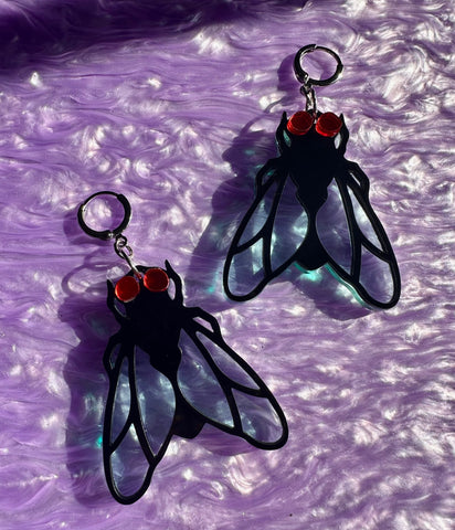 Laser-cut acrylic dangle earrings in the shape of a pair of house flies with black bodies and transparent matte finish iridescent wings and red mirrored eyes. Attaches to ears with silver metal huggie hoops. Shown on marbled purple background