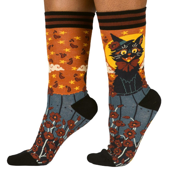 pair of unisex cotton blend crew socks with allover pattern of black witchy cat in witch hat and red bow sitting atop a wooden fence surrounded by red flowers and sky of falling red leaves and yellow stars on orange background. Shown worn by model from side