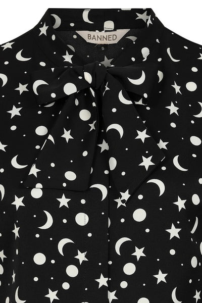 a matte finish satin long sleeve button down blouse in a black background scattered white star, crescent moon, and full moon allover pattern with a tie neck collar, slightly puffed shoulders, and button cuffed sleeves. Shown from front in close up of tie neck