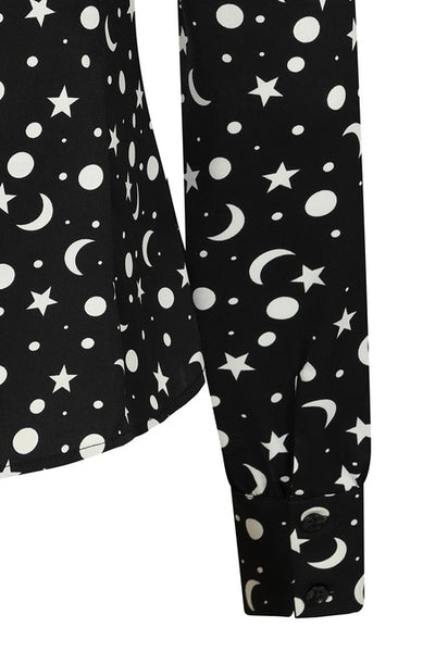 a matte finish satin long sleeve button down blouse in a black background scattered white star, crescent moon, and full moon allover pattern with a tie neck collar, slightly puffed shoulders, and button cuffed sleeves. Shown from front in close up of cuff
