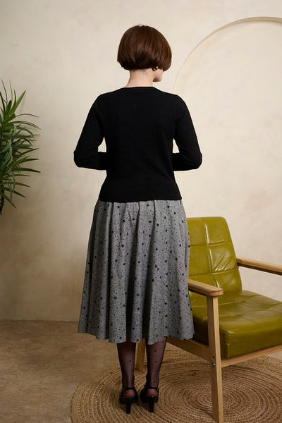 model wearing black cardigan and grey tweed high waisted swing skirt with all over flocked black star pattern, fabric covered button detail at waistband, and pockets. Shown from back
