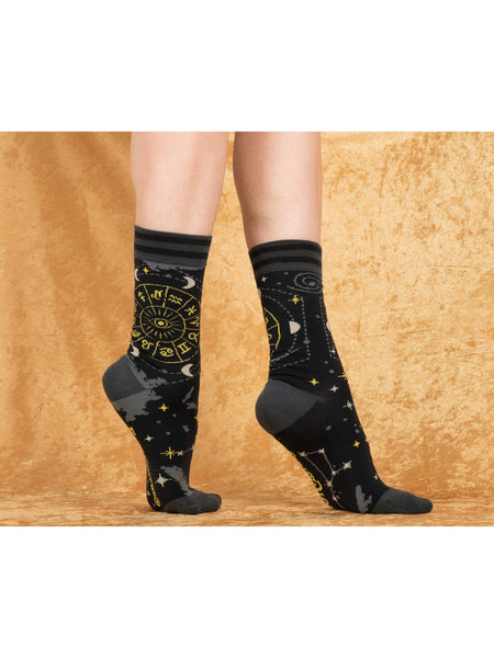Unisex crew socks with a yellow Astrology wheel illustration on the outer side of each sock and constellations, stars, moons, and clouds in grey/white/gold lurex thread as an all over pattern on a black background. Shown worn by a model in front of a gold background 