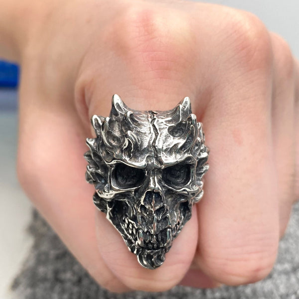 A stainless steel ring with antiqued detail in the shape of a demonic skull with horn-like features and grinning. Shown worn on a pointer finger in a fist