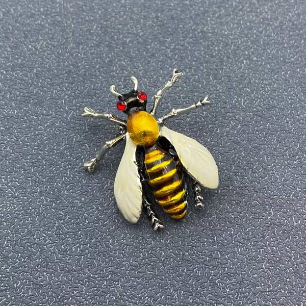 silver metal honeybee brooch with enameled white wings, black and yellow enameled thorax and abdomen, and small red jeweled eyes. Shown from front