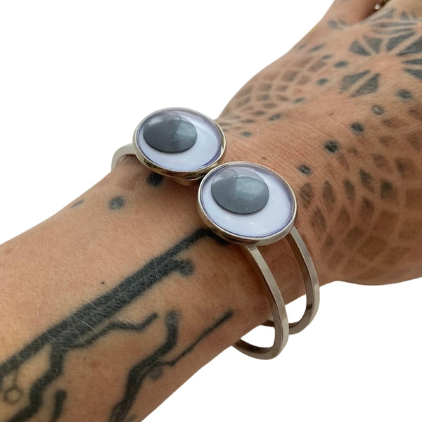 Silver metal hinged bracelet with large two black and white plastic googly eye adornments on front. Shown worn by model