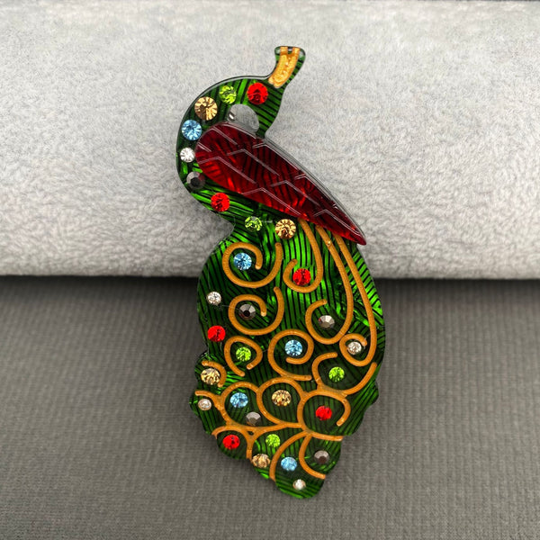 layered acrylic resin brooch of peacock in marbled dark red and green with multicolored rhinestone jewels and painted gold detail. Shown from front