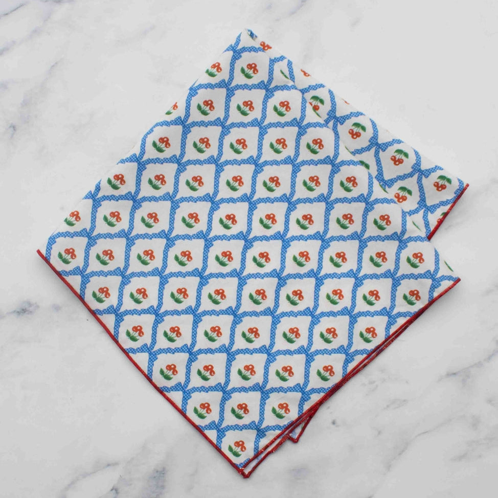 Cotton square scarf with allover blue and white gingham bow pattern & tiny red and green cherry print. Edges of scarf have red hem edge. Shown folded