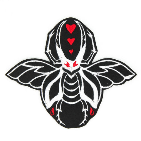 An embroidered patch of a large white beetle with wings on a black background. It has red eyes and there are three small red hearts in between its pincers. Shown flat