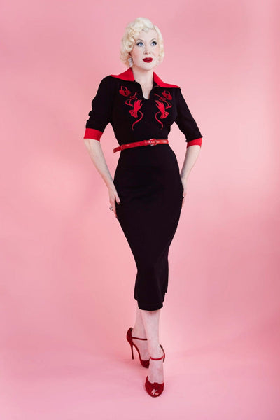 model wearing black sweater knit dress with bright red rolled collar, v-neckline, elbow length sleeves with red cuffs, and embroidered detail across the bodice in matching red of heart-shaped lock, key, and gloved hand. Dress has pencil skirt with side slit and red patent faux leather skinny belt. Shown from front