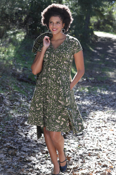 A woman wearing a stretchy cotton knit cap-sleeve olive green dress with a beige black and white woodcut style floral pattern, with faux wrap surplice bodice and notched collar v-neckline. Shown standing outdoors on a gravel path with trees in the background.
