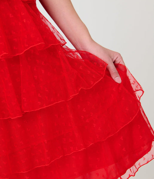 A woman modeling a bright red heart-patterned lace dress with high banded neckline, ruffled yoke bodice, and just below the knee five tiered matching lace skirt. Shown from front in close up of lace pattern on skirt
