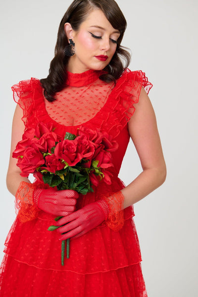 A woman modeling a bright red heart-patterned lace dress with high banded neckline, ruffled yoke bodice, and just below the knee five tiered matching lace skirt. Shown from front in close up holding bouquet of red roses