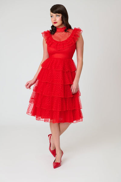 A woman modeling a bright red heart-patterned lace dress with high banded neckline, ruffled yoke bodice, and just below the knee five tiered matching lace skirt. Shown from front.