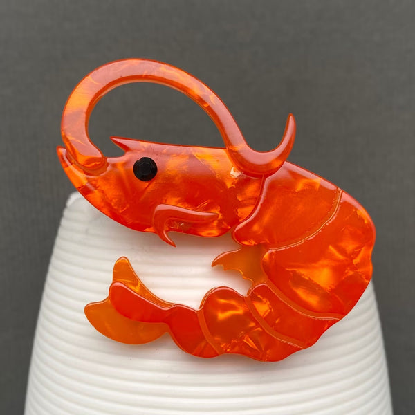 layered acrylic resin brooch of an orange marbled shrimp seen from the side with single black rhinestone jewel for an eye. Shown flat