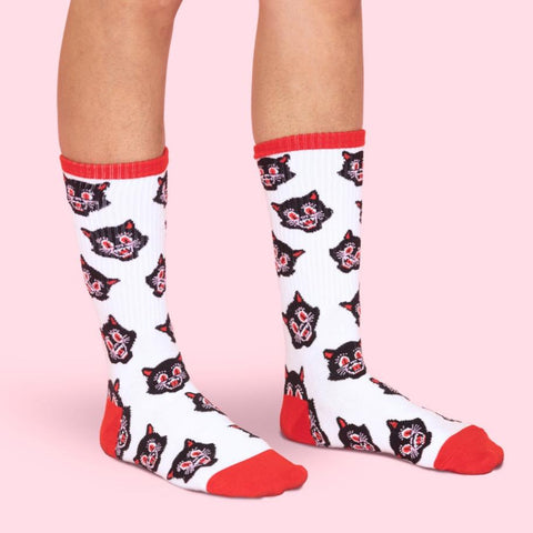 white unisex crew socks with red cuffs and toes & an all-over pattern of a black cat head with red eyes and open mouth. Shown worn by model