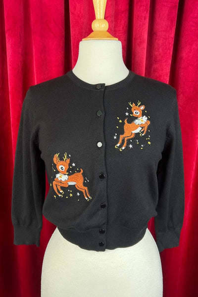 black knit 3/4 sleeve cardigan with black plastic buttons and mirrored embroidered image of reindeer leaping wearing white ruffled collar alongside yellow and white stars, on upper left bodice and bottom right. Shown on dress form