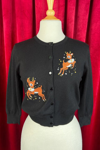 black knit 3/4 sleeve cardigan with black plastic buttons and mirrored embroidered image of reindeer leaping wearing white ruffled collar alongside yellow and white stars, on upper left bodice and bottom right. Shown on dress form