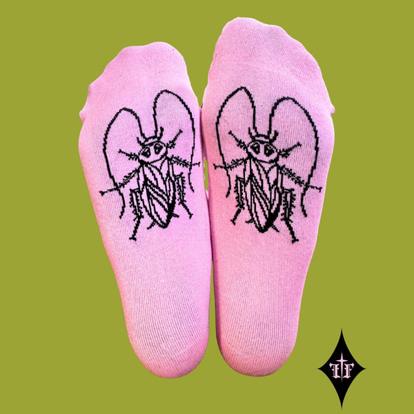 Bright pink cotton-poly blend crew socks with lettuce hem-style ruffled ribbed cuffs and “Filthy” written in black script running across each outer cuff and black outline cockroach on bottom of sock. Shown worn by model shown from bottom of feet to display both cockroaches on green background 