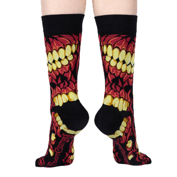 A pair of unisex cotton blend crew socks with a black background and an allover pattern of a skull's teeth and mouth in red and pale yellow. Shown worn by model from back to display black heels